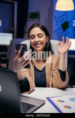Saying hello in the course of teleconference with entrepreneurs doing overtime. Woman working on finance during a video conference with coworkers at night hours in the office. Stock Photo