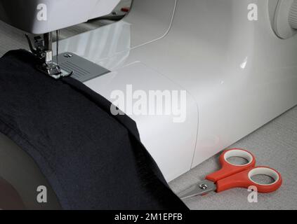 Home sewing machine at work. Close-up of the mechanism, a piece of black cloth, red scissors on the table. Stock Photo