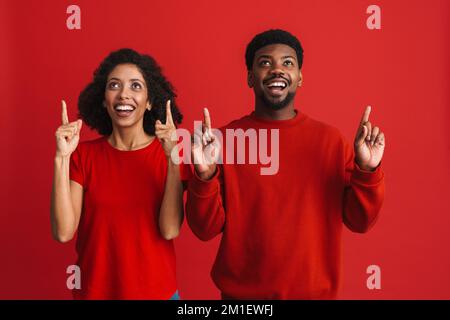 Black happy man and woman smiling and pointing upward at copyspace isolated over red background Stock Photo