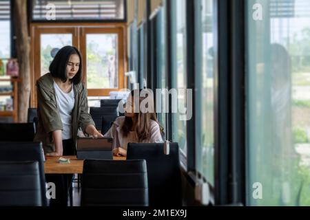 The two women discussing a topic while using a tablet at a table in the coffee shop Stock Photo
