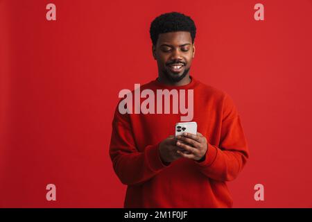 Black bearded young man smiling and using cellphone isolated over red background Stock Photo