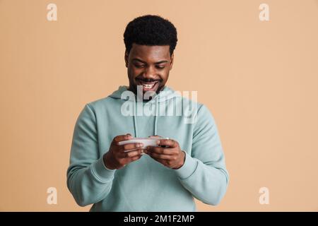Black bearded young man smiling and using cellphone isolated over beige background Stock Photo