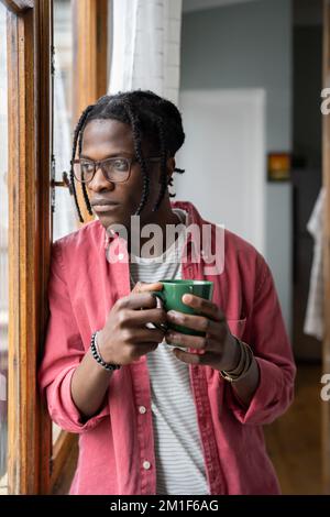Depressed sad African guy holding cup of tea looking out window, having problems in personal life Stock Photo
