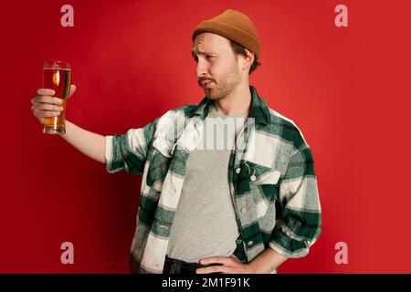 Portrait of young man in casual checkered shirt posing with beer isolated over red background. Mates Stock Photo