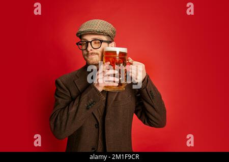 Portrait of stylish man in classical clothes posing with glass of foamy lager beer isolated on red background. Friday evening refreshment Stock Photo