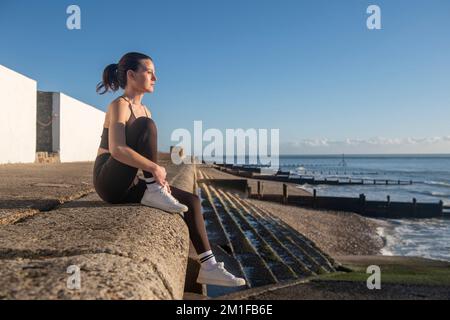 Sporty woman sitting on a wall by the sea resting after exercising outside. Enjoying the sun and the view. Stock Photo