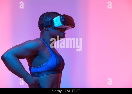 Happy young black lady in VR glasses exploring cyberspace, touching imaginary screen, playing virtual reality game in neon light. Positive African Ame Stock Photo