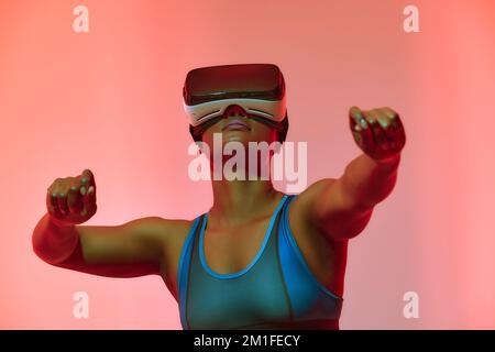 Happy young black lady in VR glasses exploring cyberspace, touching imaginary screen, playing virtual reality game in neon light. Positive African Ame Stock Photo