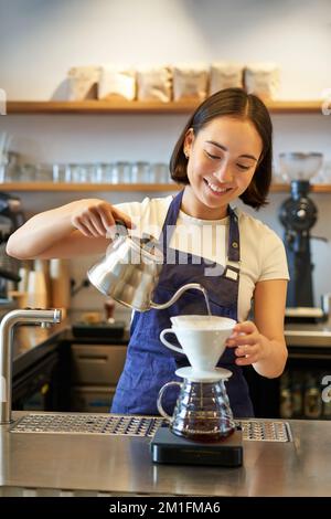 Vertical shot of smiling asian bartender, barista in blue apron, pouring water with small kettle, brewing coffee behind counter in her cafe Stock Photo