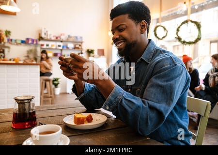 Black smiling man using mobile phone while having brunch in cafe indoors Stock Photo