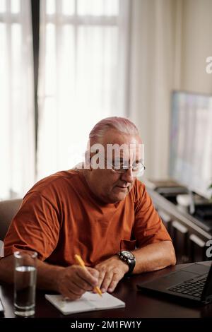 Senior man in glasses work g on laptop at home and taking notes in planner Stock Photo