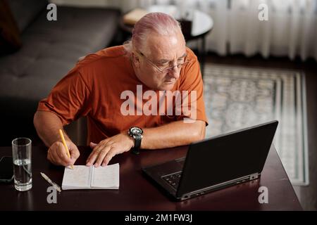Senior man watching educational video on laptop and taking notes in notebook Stock Photo