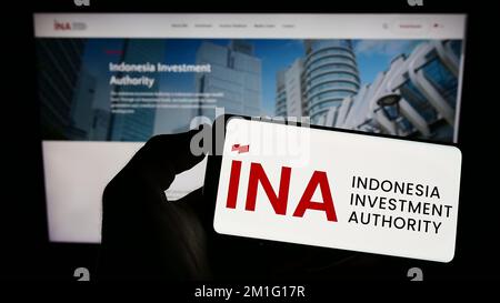 Person holding mobile phone with logo of Indonesia Investment Authority (INA) on screen in front of business web page. Focus on phone display. Stock Photo