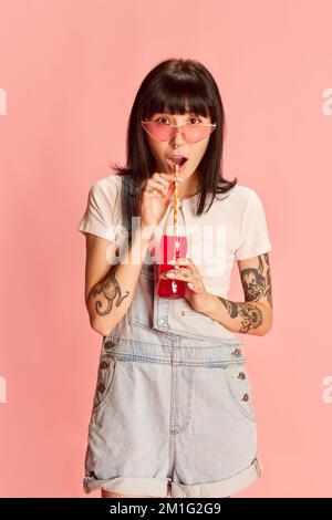 Young stylish girl with tattoos posing in denim overalls, drinking soda isolated on pink background Stock Photo