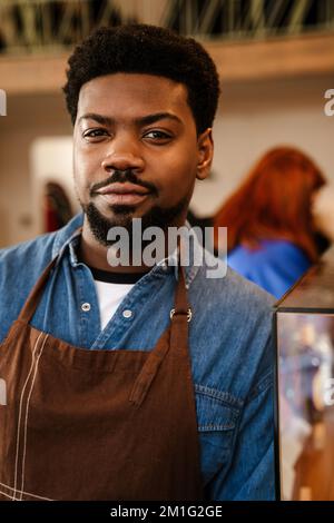 Black bearded man wearing apron standing while working in cafe indoors Stock Photo