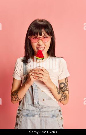 Young stylish girl in trendy sunglasses, with tattoos, posing in denim overalls, eating lollipop isolated on pink background Stock Photo