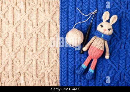 Knitted children's toy hare or bunny with a balls of thread and a crochet hook on a knitted blue and beige cloth. The symbol of 2023. Stock Photo