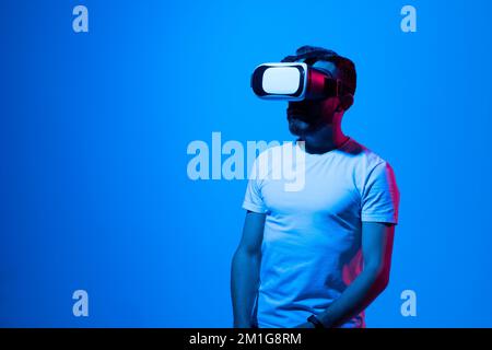 Bearded brunette man in white t-shirt play VR games in virtual reality helmets in neon colourful light. Guy in VR helmets play video games. VR gaming Stock Photo