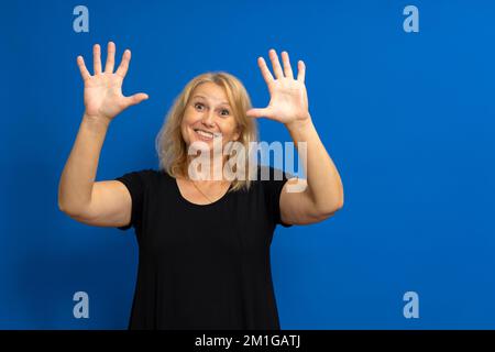 Pretty caucasian blonde woman in her 40s dressed in a black t-shirt raising her palms up while smiling at the camera, isolated on blue studio Stock Photo