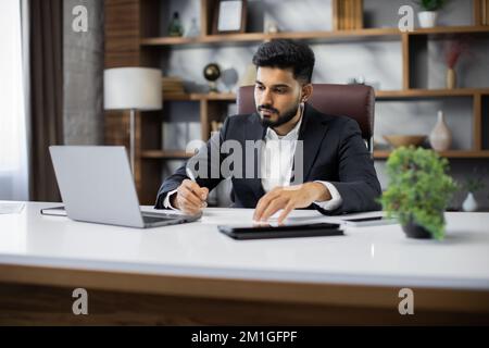 Confident businessman wearing suit writing notes or financial report, sitting at desk with laptop, focused serious man working with paper documents, student studying online, research work Stock Photo