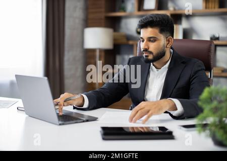 Confident businessman wearing suit writing notes or financial report, sitting at desk with laptop, focused serious man working with paper documents, student studying online, research work Stock Photo
