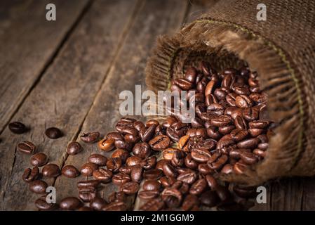 Sack of coffee beans overturned on an old rustic table with the beans scattered on top. Stock Photo