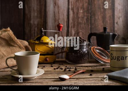Cup of fresh coffee next to a wooden spoon with sugar, in the background an old coffee grinder, an Italian coffee maker and a jar with coffee. Stock Photo