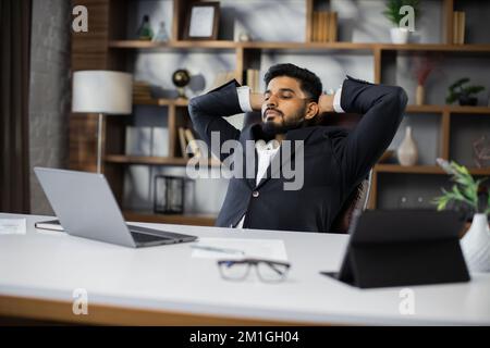 Confident man in glasses sitting relax at home office workplace take nap or daydream. Happy relaxed Caucasian young male rest in chair distracted from computer work, relieve negative emotions. Stock Photo