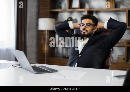 Confident man in glasses sitting relax at home office workplace take nap or daydream. Happy relaxed Caucasian young male rest in chair distracted from computer work, relieve negative emotions. Stock Photo