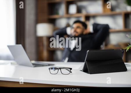 Focus on eye glasses, tablet and laptop. Happy relaxed caucasian young male rest in chair distracted from computer work, relieve negative emotions. Desktop background, copy space. Stock Photo