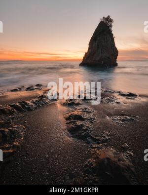 A vertical long exposure shot of sea waves breaking on the shore with a rock in the backdrop at sunset Stock Photo