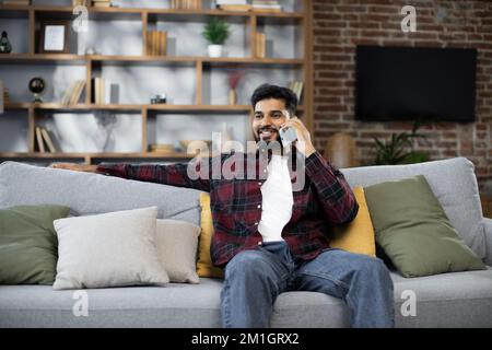 Handsome bearded positive man sitting on couch and calling to his friend using smartphone indoors. Smiling male freelancer taking break during working process at home. Stock Photo