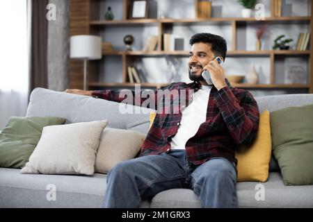Handsome bearded positive man sitting on couch and calling to his friend using smartphone indoors. Smiling male freelancer taking break during working process at home. Stock Photo