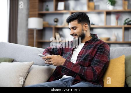 Attractive man in red checkered shirt sitting on couch and using modern smartphone indoors. Male freelancer taking break during working process at home. Stock Photo