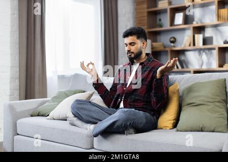 Yong male sitting on couch in lotus pose put hands on lap folded fingers closed eyes enjoy meditation do yoga exercise at home. No stress healthy habit mindfulness lifestyle anxiety relief concept Stock Photo
