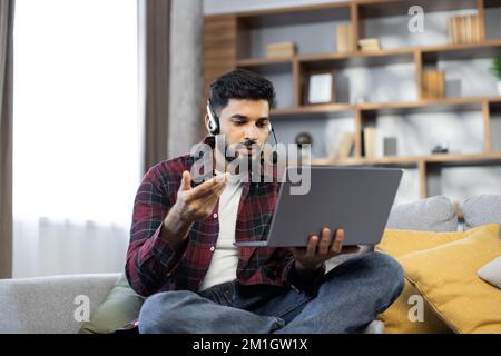 Closeup of young student man in wireless headset watching online lesson on laptop, studying foreign language, attending virtual class, webinar, talking on video call to teacher, resting on couch Stock Photo
