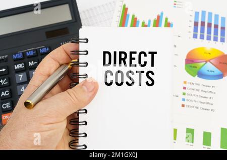 Finance and economics concept. The man is holding a pen and a notebook with the inscription - DIRECT COSTS. In the background are financial charts, ch Stock Photo