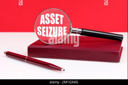 Business and finance concept. On a white and red background, a notebook, a pen and a magnifying glass, inside which the inscription - Asset Seizure Stock Photo