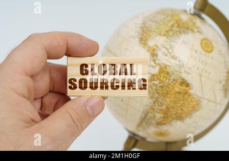Globalization concept. A man holds in his hand a wooden plate on which it is written - GLOBAL SOURCING. In the background is a globe. Stock Photo
