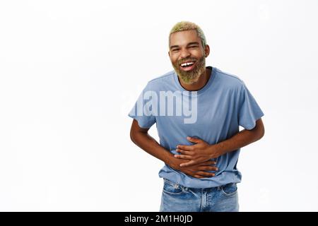 Handsome hipster guy laughs and smiles, holds hands on belly as he chuckles, stands over white background Stock Photo