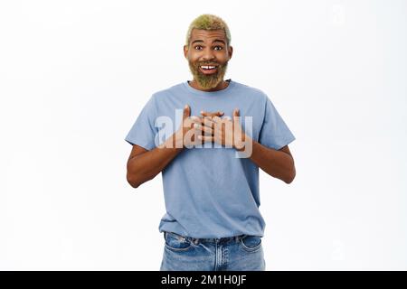 Portrait of surprised happy guy, looks with disbelief and appreciation, holds hands on chest, flick eyebrows amazed, stands over white background Stock Photo