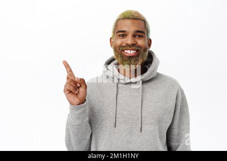 Portrait of bearded hipster guy, yellow hairstyle and long beard, smiling and pointing left at banner, showing advertisement space Stock Photo