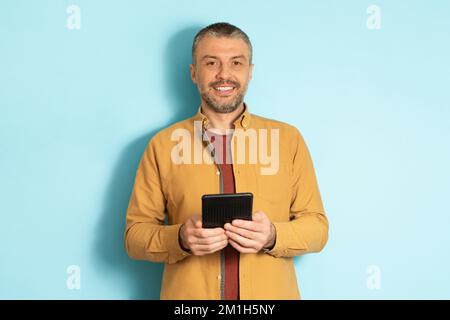 People and technology concept. Portrait of mature caucasian man holding and using tablet standing over blue background Stock Photo