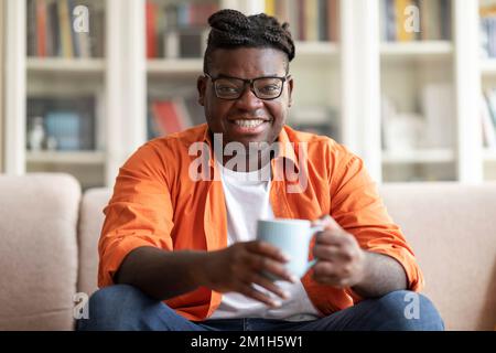 Smiling black guy sitting on couch with mug of tea Stock Photo