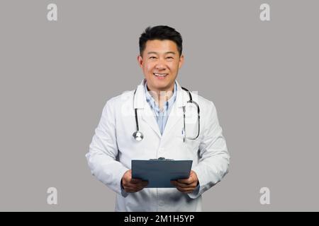 Smiling mature chinese man therapist in white coat with stethoscope use tablet, isolated on gray background Stock Photo