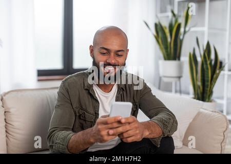 Handsome latin man using smartphone while resting on sofa at home, browsing internet or messaging with friends Stock Photo