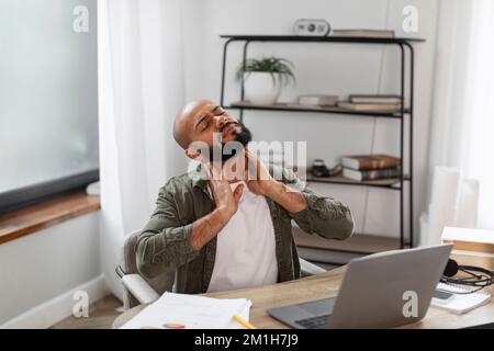 Upset latin male freelancer sitting at workplace at home, using laptop and rubbing his neck with both hands Stock Photo