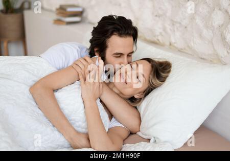 Loving caucasian couple relaxing in bed at home, enjoying togetherness and tenderness in love, man kissing wife Stock Photo
