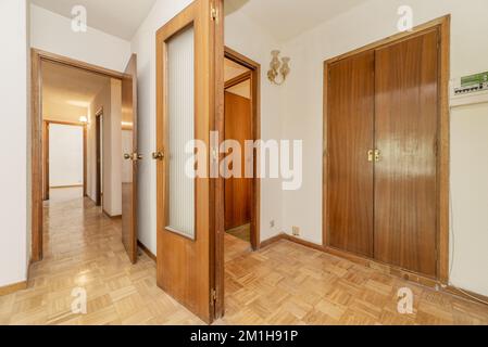 A room with built-in wooden wardrobes next to a corridor with a wooden and glass door and a checkerboard oak floor Stock Photo