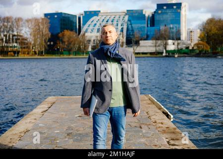 Portrait of middle-aged man standing on concrete pier at river in city in autumn, looking at camera, holding laptop. Stock Photo